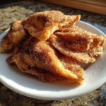 A pile of freshly fried buñuelos coated heavily in cinnamon sugar, resting on a white plate.