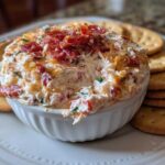 A white bowl filled with creamy, cheesy hoagie dip topped with bacon bits, served with round crackers.