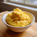 A close-up of a white bowl filled with fluffy, bright yellow, creamy cheese grits sitting on a wooden surface.