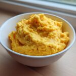 A white bowl filled with fluffy, bright yellow, creamy cheese grits sitting on a wooden surface near a window.