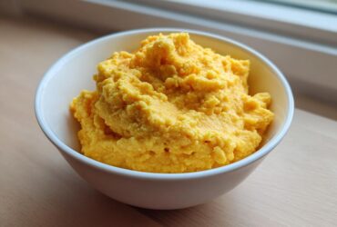 A white bowl filled with fluffy, bright yellow, creamy cheese grits sitting on a wooden surface near a window.