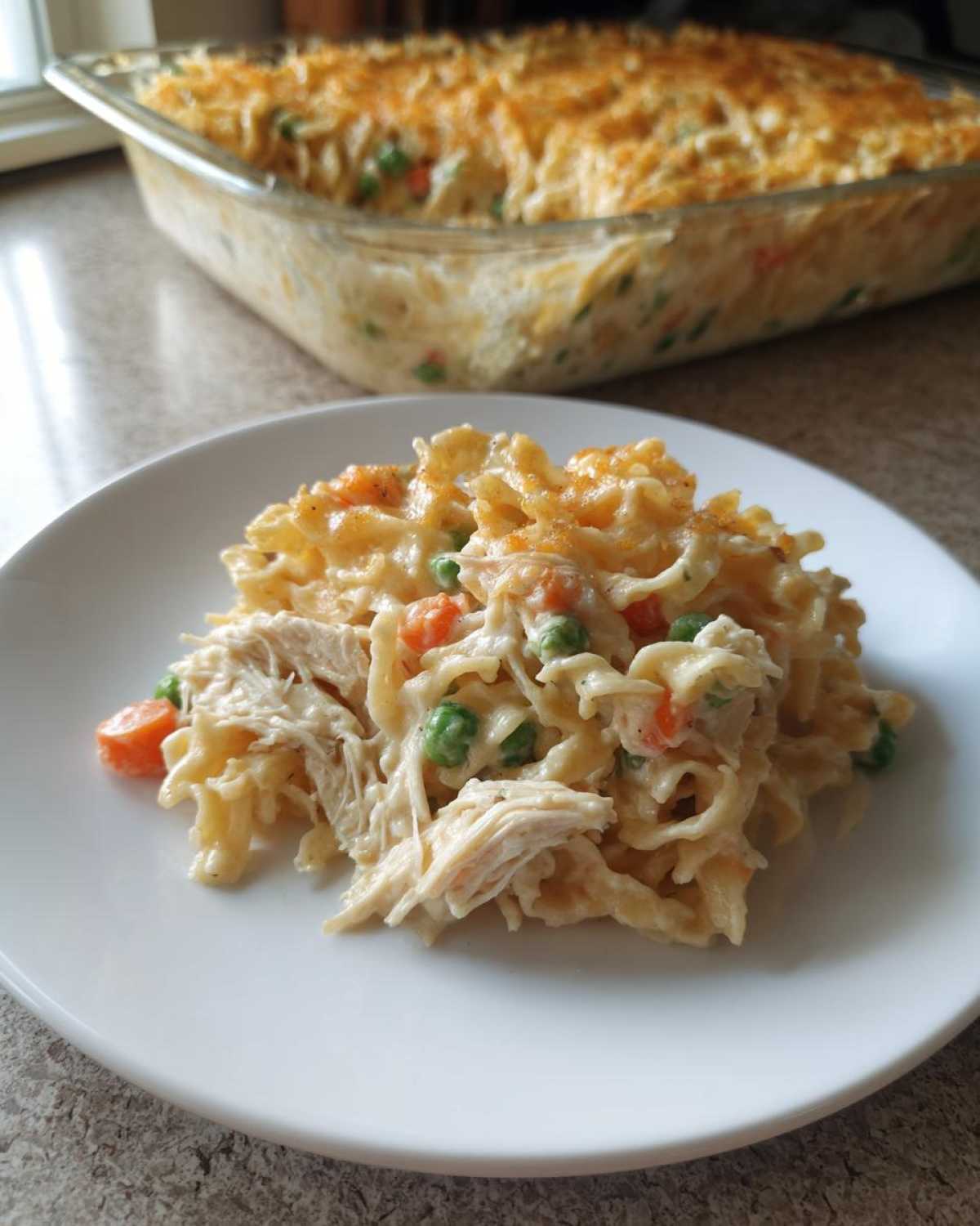 A serving of creamy chicken noodle casserole with shredded chicken, peas, and carrots on a white plate, with the full dish in the background.