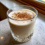 A glass of creamy, light brown coquito topped with a sprinkle of ground cinnamon.