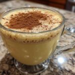 Close-up of a glass mug filled with creamy, yellow eggnog, topped with a dusting of ground cinnamon.