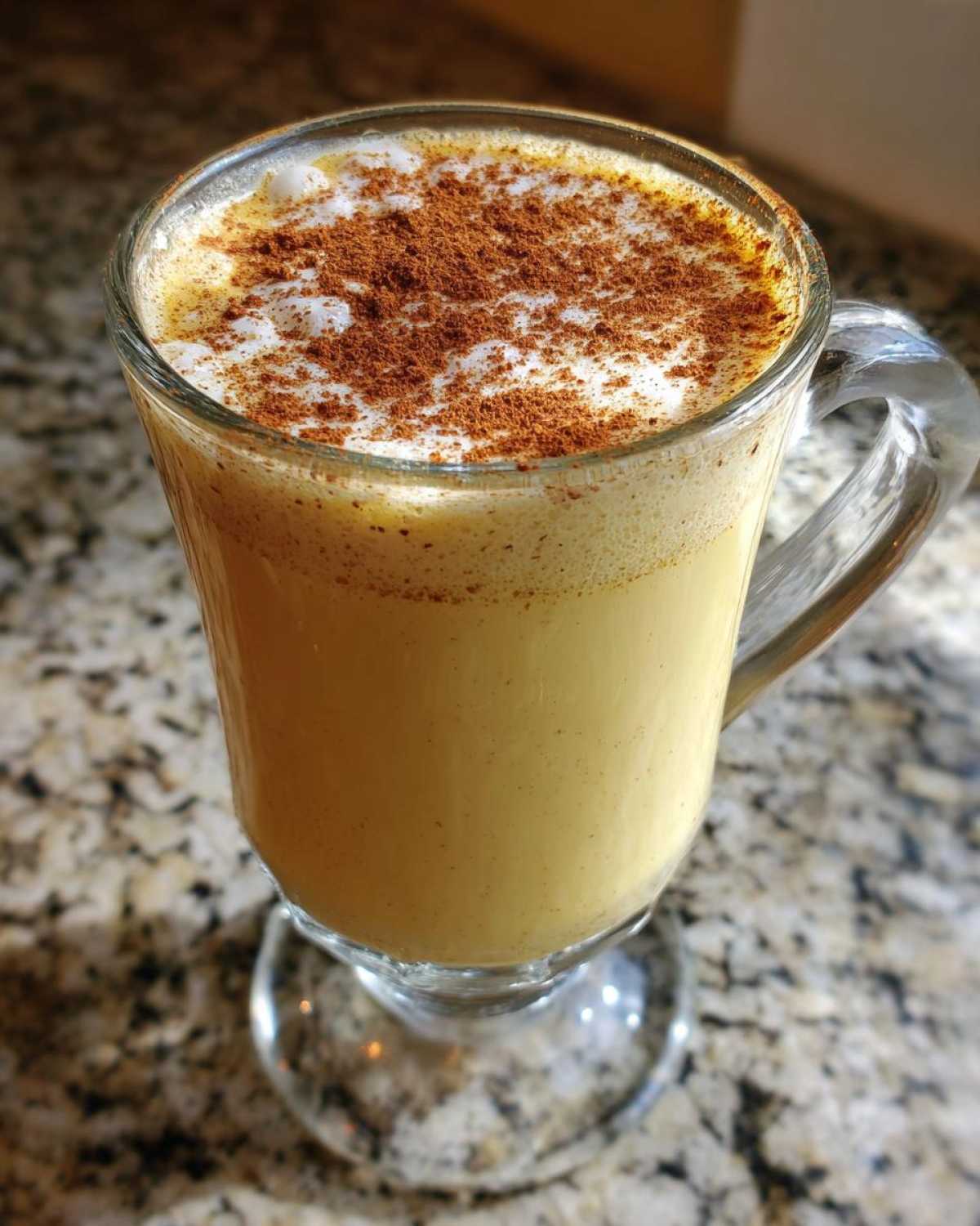 Close-up of a glass mug filled with creamy yellow eggnog, topped with foam and a generous dusting of cinnamon.