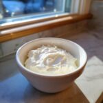 A white bowl filled with thick, white creamy horseradish sauce sitting on a countertop near a window.
