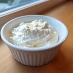 A white, fluted ramekin filled high with thick, white creamy horseradish sauce, sitting on a wooden surface near a window.