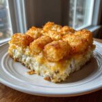 A square serving of creamy tater tot casserole topped with crispy, golden tater tots, served on a white plate.