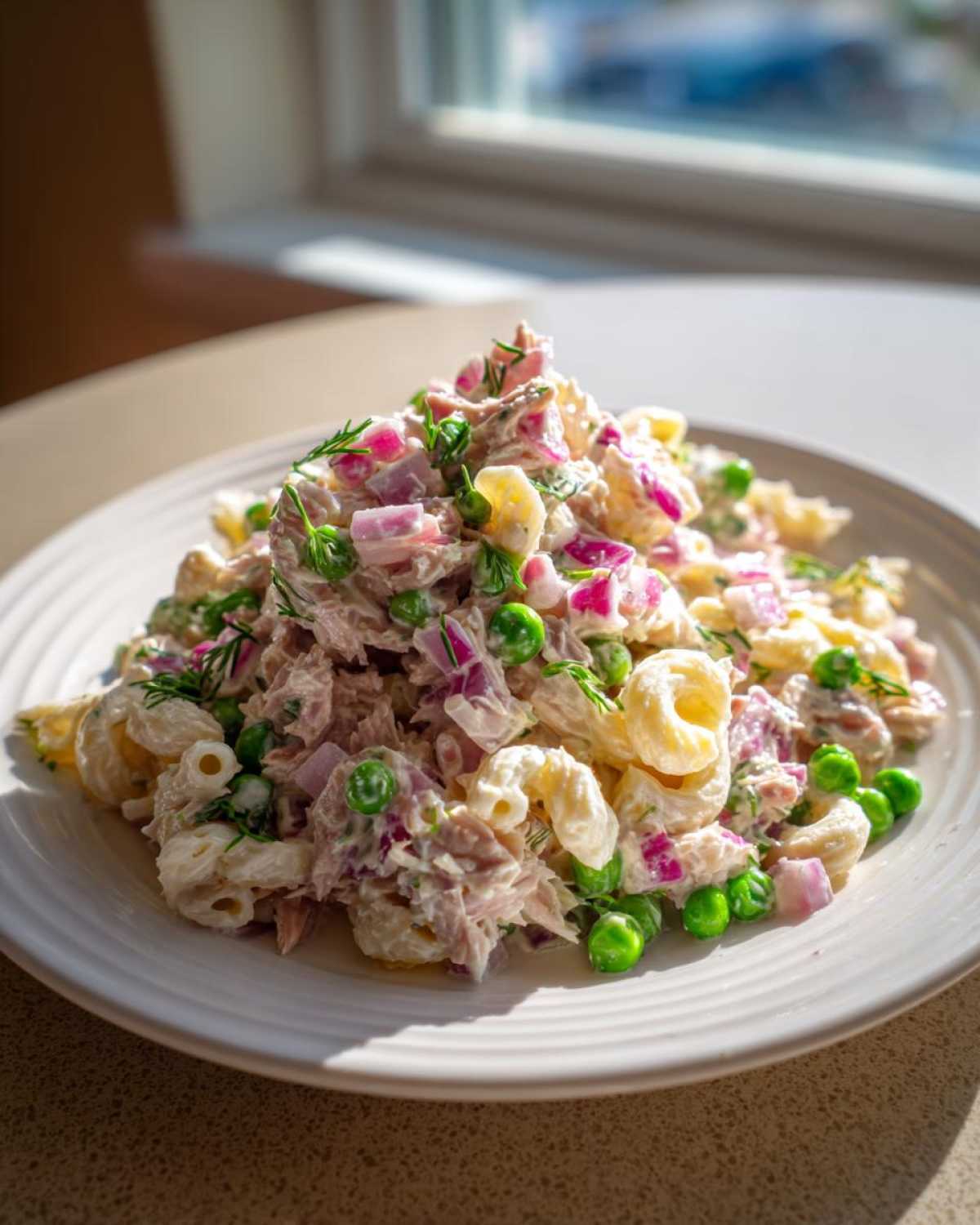 A generous serving of creamy tuna pasta salad mixed with bright green peas, diced red onion, and fresh dill, served on a white plate.