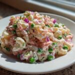 A close-up of creamy Tuna Pasta Salad mixed with elbow macaroni, green peas, red onion, and herbs on a white plate.