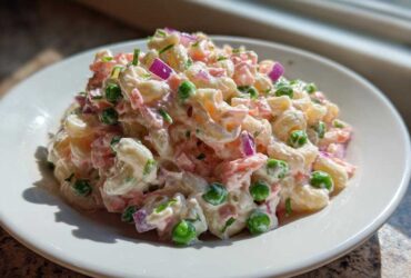 A close-up of creamy Tuna Pasta Salad mixed with elbow macaroni, green peas, red onion, and herbs on a white plate.