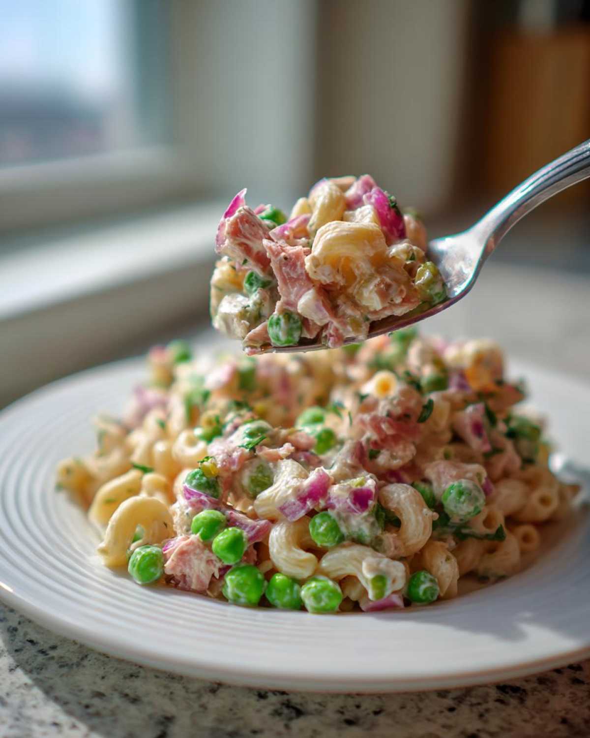 A fork lifts a bite of creamy Tuna Pasta Salad mixed with bright green peas and red onion over a plate of the salad.