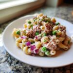 A close-up of a serving of creamy Tuna pasta salad mixed with rotini pasta, bright green peas, and diced red onion, garnished with parsley.