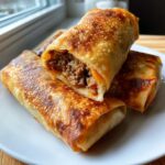 Close-up of crispy, golden-brown Chimichangas stacked on a white plate, one cut open showing the seasoned ground beef filling.