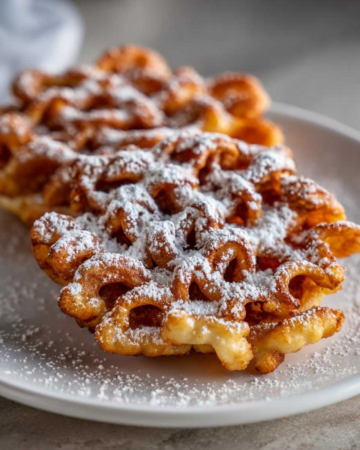 Close-up of a golden brown, crispy funnel cake recipe generously dusted with white powdered sugar on a white plate.