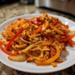 A white plate piled high with seasoned, shredded chicken mixed with sliced red and yellow bell peppers and onions, ready for crockpot chicken fajitas.