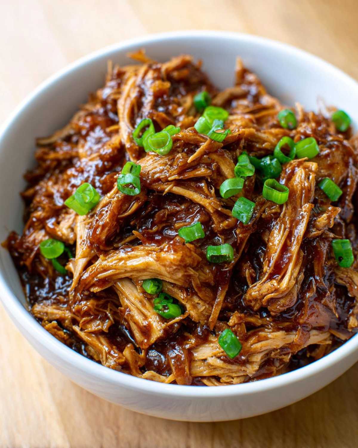 Close-up of shredded crockpot teriyaki chicken coated in rich sauce and topped with sliced green onions.