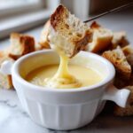 A piece of toasted bread being dipped into a small white bowl of smooth, yellow cheese fondue recipe.