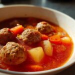 Close-up of a white bowl filled with rich, red broth albondigas soup featuring large meatballs, carrots, and potatoes.