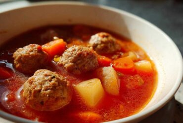 Close-up of a white bowl filled with rich, red broth albondigas soup featuring large meatballs, carrots, and potatoes.