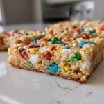 A close-up shot of a thick, chewy christmas cookie bars piece topped with colorful M&Ms and sprinkles.