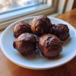 Five decadent, chocolate-coated Oreo balls stacked slightly on a small white plate near a window.