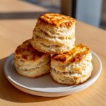Three tall, flaky buttermilk scones stacked on a light gray plate, showing golden brown tops.