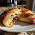 A flaky, golden brown chocolate croissant cut in half on a white plate, revealing melted chocolate filling.