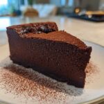 A dense, dark slice of flourless chocolate torte dusted with cocoa powder, served on a white plate.