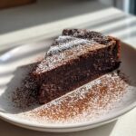 A decadent slice of flourless chocolate torte dusted heavily with powdered sugar on a white plate.