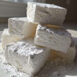 A stack of large, white, fluffy homemade marshmallows dusted heavily with powdered sugar on a white plate.