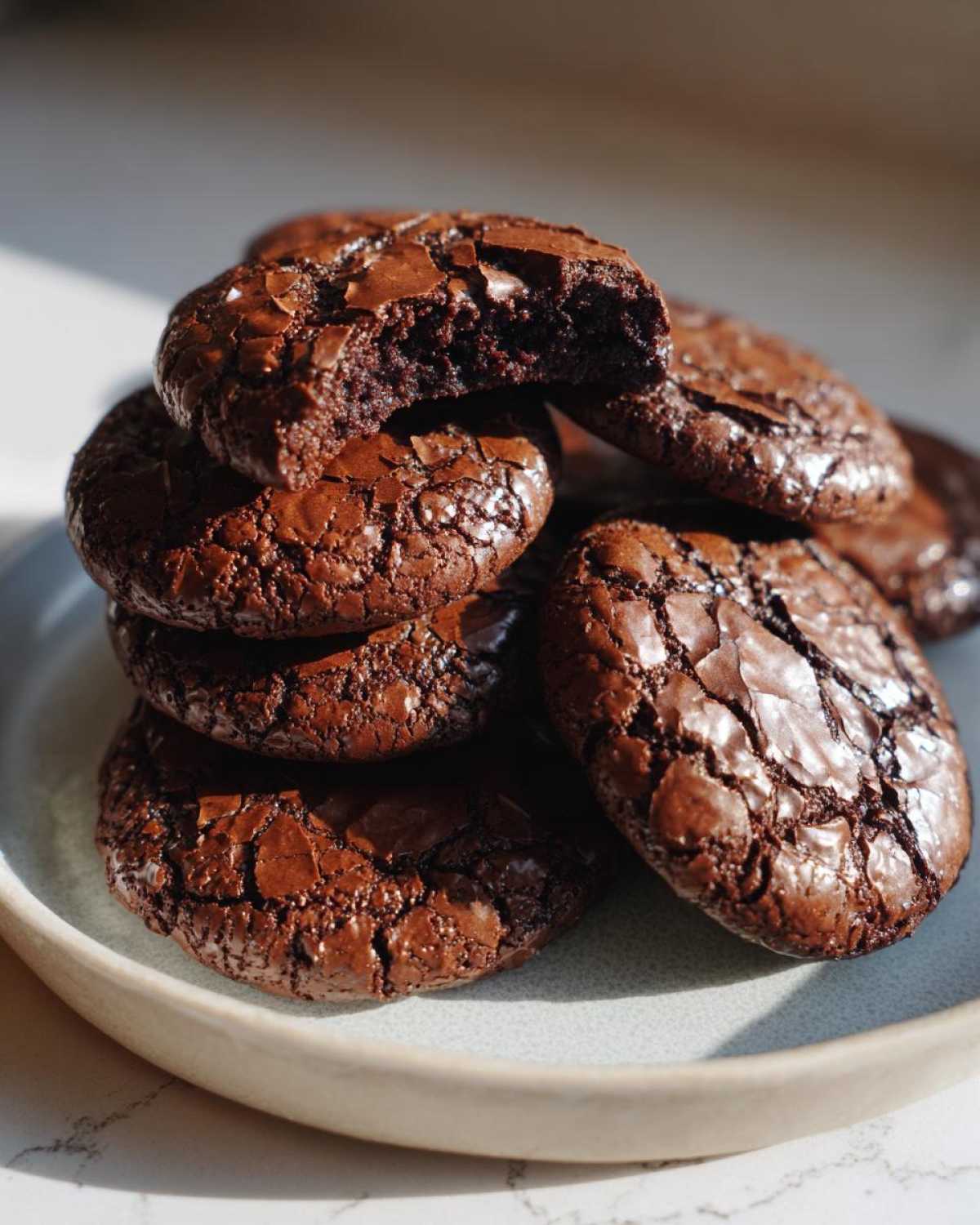 A stack of rich, dark chocolate brownie cookies with shiny, cracked tops, one cookie broken open to show the fudgy interior.