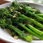 Close-up of vibrant green, sautéed broccolini tossed with browned garlic pieces on a white plate.