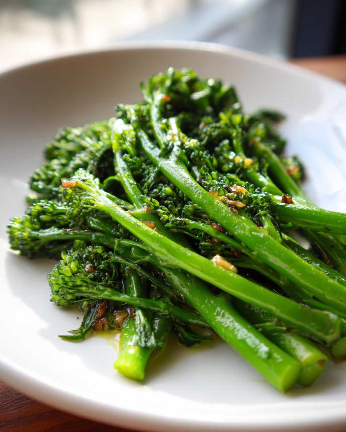 Close-up of vibrant, glossy Garlic Lemon broccolini spears lightly coated in oil and garlic bits, served on a white plate.