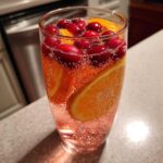 Close-up of a tall glass filled with bubbly pink champagne punch, garnished with fresh cranberries and orange slices.