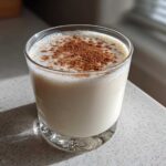 A glass filled with creamy, white coquito topped generously with ground cinnamon, sitting on a light countertop.