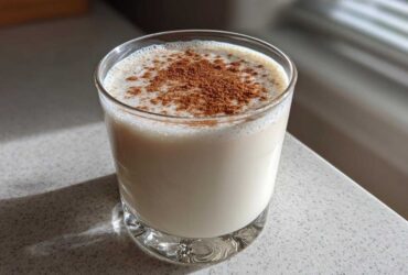 A glass filled with creamy, white coquito topped generously with ground cinnamon, sitting on a light countertop.