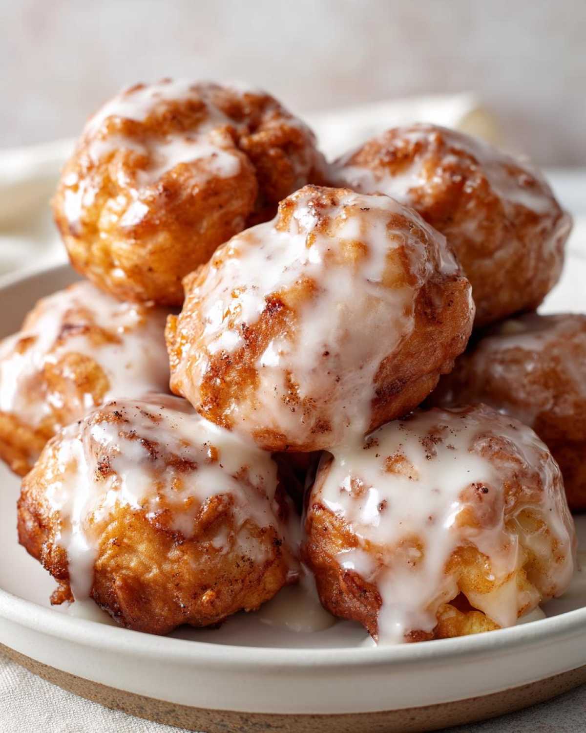 A close-up of several golden brown baked apple fritters piled high and drizzled generously with white vanilla glaze.