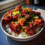 Close-up of a bowl filled with white rice topped with crispy, glazed gochujang chicken pieces and garnished with sesame seeds and green onions.