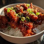 Close-up of a bowl filled with white rice topped with crispy, glazed gochujang chicken, sprinkled with sesame seeds and green onions.