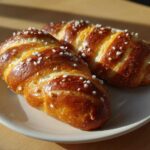Two golden brown, braided pretzel dogs dough pieces sprinkled with coarse salt on a light plate.