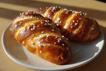 Two golden brown, braided pretzel dogs dough pieces sprinkled with coarse salt on a light plate.