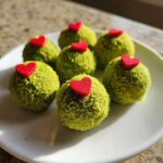 Six bright green Grinch Oreo balls coated in sprinkles, each topped with a small red heart candy on a white plate.