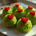 Six bright green Grinch Oreo Balls coated in sprinkles, each topped with a small red, sugary heart candy.