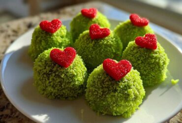 Six bright green Grinch Oreo Balls coated in sprinkles, each topped with a small red, sugary heart candy.