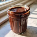 A glass jar filled with thick, homemade chocolate syrup, dripping down the side in bright sunlight.