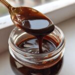 A golden spoon holds thick, glossy chocolate syrup dripping back into a clear glass jar.