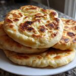 A stack of four fluffy, golden-brown homemade pita bread rounds resting on a white plate.