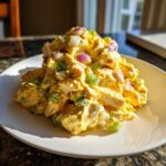 A mound of creamy, yellow honey mustard chicken salad mixed with red onion and green onions on a white plate.