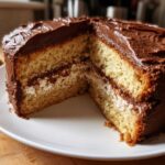 Close-up of a slice removed from an Italian Love Cake showing two layers of light sponge cake, a creamy white filling, and rich chocolate frosting.
